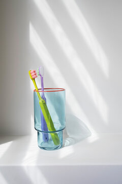 Two colorful toothbrushes stand in a glass on a white shelf