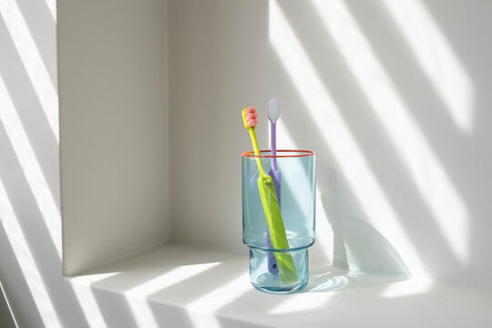 Two colorful toothbrushes stand in a glass on a white shelf