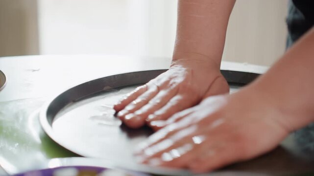 Close-up of adult hands pressing and smoothing round dough on dark nonstick metal baking tray over glossy kitchen countertop, creating even thickness with gentle downward pressure and careful