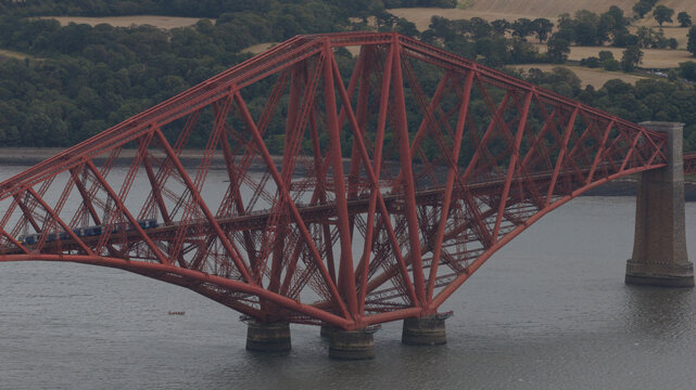 Aerial view of the iconic red Forth Bridge spanning the Firth of Forth with a train crossing the tracks, surrounded by lush green hills in Queensferry, United Kingdom.