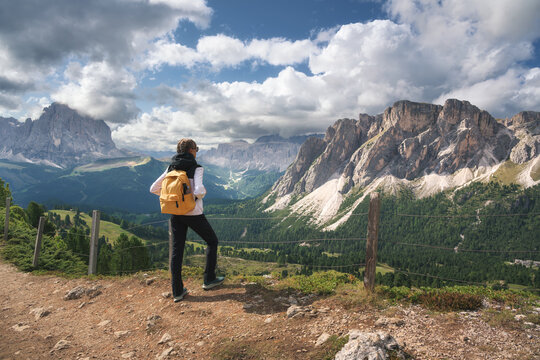 Woman hiker with yellow backpack standing on trail looking at mountain range in Dolomites, Italy. Alpine summer landscape with female tourist enjoying valley view, rocky peaks and cloudy sky. Hiking