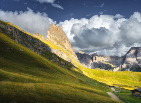 Seceda mountain peak with green grass meadow slope under dramatic cloudy sky at sunset in Dolomites, Italy. Alpine autumn landscape with rocky cliffs, sunbeams, wooden hut and hiking path. Travel