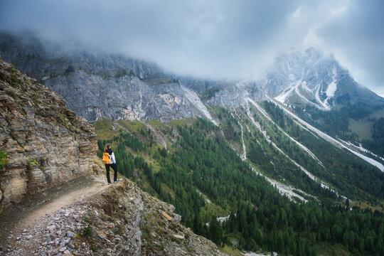 Woman hiker with yellow backpack standing on cliff trail looking at mountain range in Dolomites, Italy. High altitude alpine landscape with girl, rocky peaks, fog, green forest, overcast dramatic sky