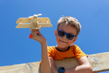 A happy boy climbed a high fence and held a wooden airplane in his hand against the blue sky. © Prrrettty
