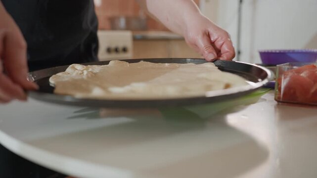 Close-up of hands guiding, pressing, and smoothing thin dough into circular metal tray on white countertop; visible motion of stretching and tucking edges creates taut, translucent dough surface