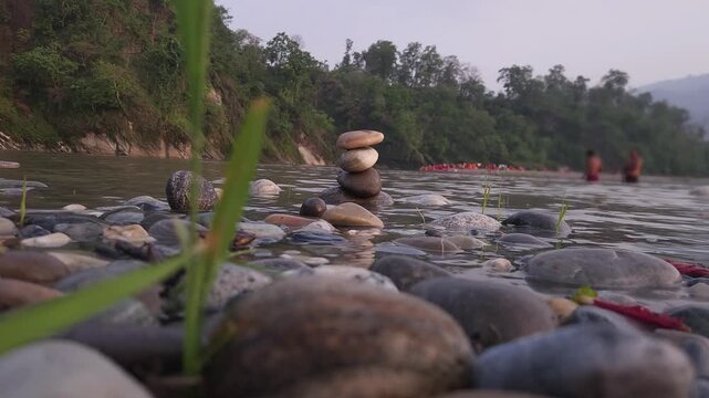 Defocused Sacred River Bathing at Devghat Dham, Nepal, with Balanced River Stones in Foreground, Hindu Pilgrimage, Spiritual Ritual, Peaceful Riverside Landscape and Cultural Heritage