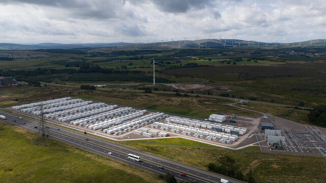 Aerial view of the BESS battery energy storage facility with rows of white units, near a highway and surrounded by rolling hills and wind turbines in Lanark, Scotland, United Kingdom.