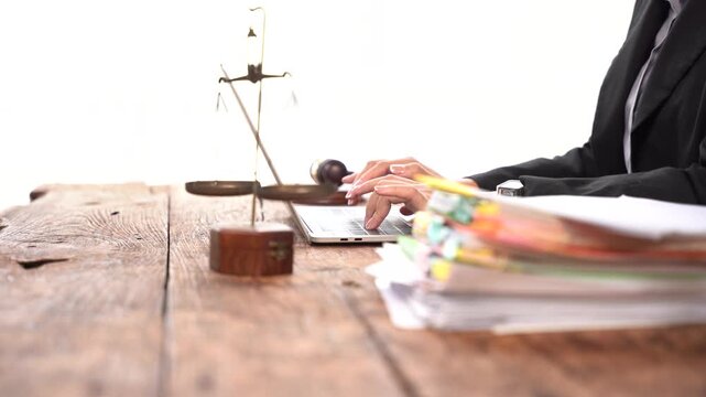 Legal Professional Working: A legal professional's hands are seen typing on a laptop next to a set of scales of justice and a stack of documents, symbolizing the pursuit of justice and legal work.