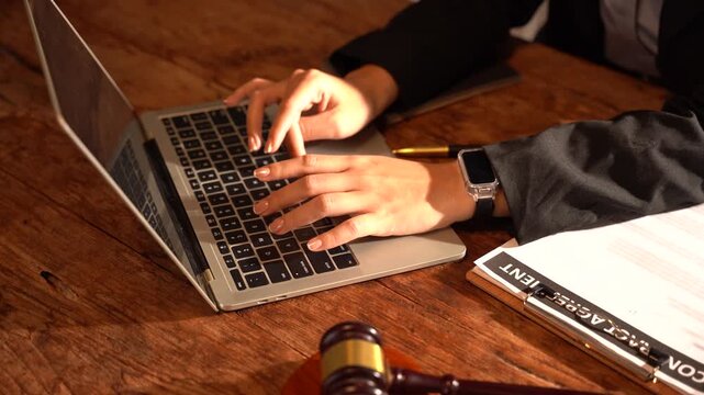Legal Professional at Work: A legal professional's hands type diligently on a laptop, with a gavel and legal documents on a wooden desk, symbolizing justice and legal practice.