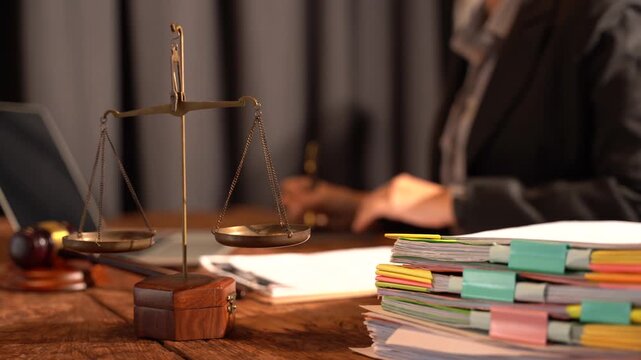 Justice Embodied: Scales of justice, a gavel, and a stack of legal documents on a wooden desk, with a blurred legal professional in the background, symbolize the essence of law and order.