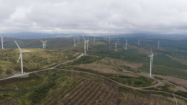 Aerial view of a wind farm with multiple white wind turbines situated on rolling green hills under a cloudy sky in Lanark, Scotland, United Kingdom.