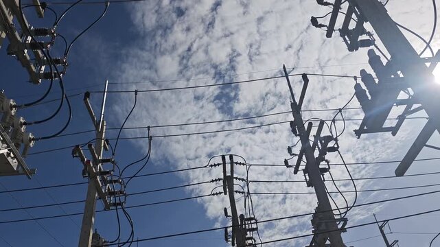 Pan right video of automatic circuit and electrical power distribution equipment on concrete poles under dramatic cloudy sky with bright sunlight.
