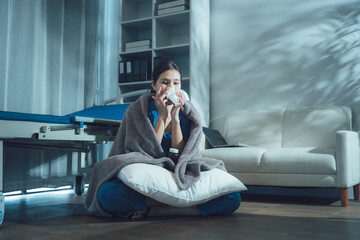 Feeling Unwell: A young woman wrapped in a cozy blanket sits on the floor, holding a tissue and looking unwell. The dim lighting and the room setting suggest a feeling of sickness and discomfort. © Kritdanai