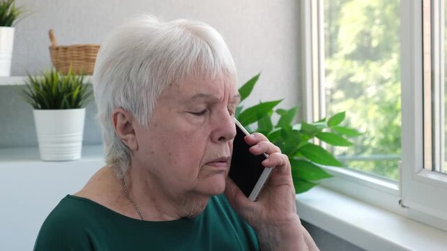 Serious elderly woman talking on mobile phone at home close up. Senior lady refuses something, not confirm information from scammer. Concept of scam or spam call