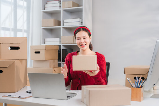 Small Business Owner Unboxing: A cheerful young woman, a small business owner, unboxes a package while engaging with customers online, surrounded by her inventory of goods ready for shipping.