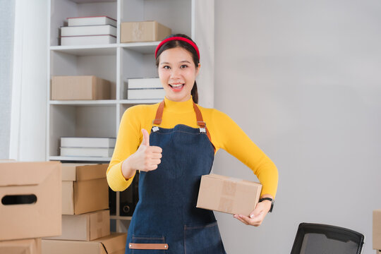 Happy Entrepreneur with Package: A smiling female entrepreneur in an apron gives a thumbs up, holding a small package, surrounded by boxes in a storage area. Ready for shipping!
