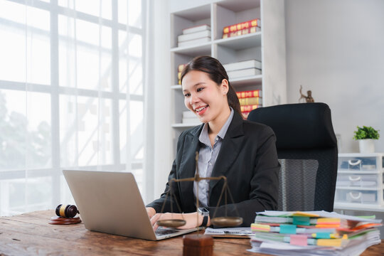 A Female Lawyer Works From Her Office: A focused and smiling young female lawyer in a suit sits at her desk in a bright, well-organized office.