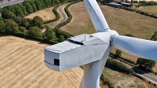 Aerial view of Penny Hill Wind Farm featuring a large white wind turbine nacelle and blades rising above golden agricultural fields and a nearby road in Aston, England, United Kingdom.
