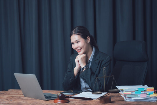 Lawyer at Work: A focused female lawyer in a sharp suit beams at her laptop screen, her hands clasped thoughtfully before her. A gavel and stack of documents sit on her desk.