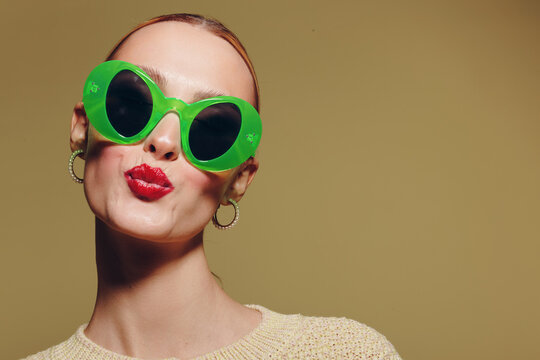 Young woman with bright red lipstick wearing large green sunglasses and hoop earrings poses with puckered lips on beige background. Concept of fashion, style, and playful attitude.