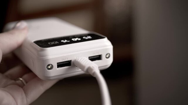 A close-up of a female hand holding a white portable power bank with a white USB cable plugged in. Concept of mobile energy, external battery backup, and digital lifestyle on the go.