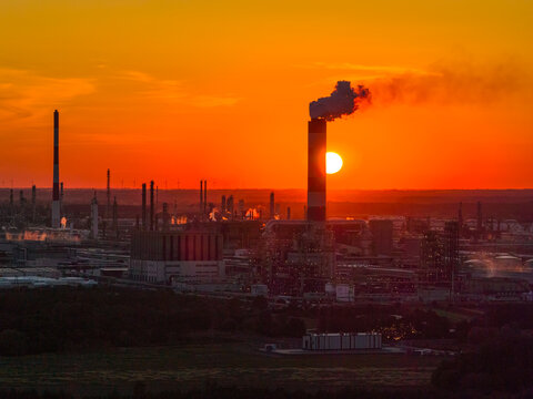 Aerial view of the PKN Orlen refinery at sunset, featuring a chimney emitting smoke against the orange sky, with industrial infrastructure in Plock, Masovian Voivodeship, Poland.