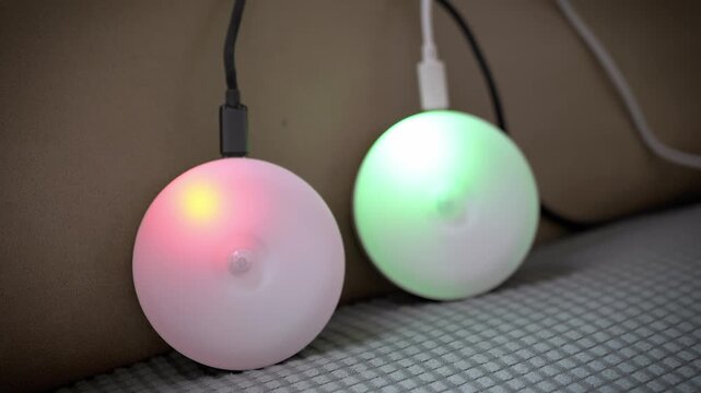 Two round wireless motion sensor night lights charge with USB cables on a textured couch. Concept of smart home technology, battery charging, energy, and modern household gadgets.