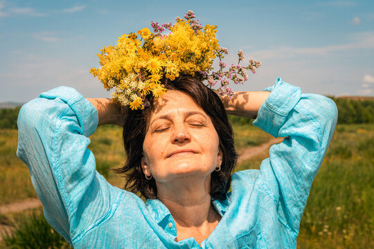 Woman with closed eyes and wreath of wildflowers on her head, wearing turquoise linen shirt, shot low against blue sky with light clouds. Women's mental health, slow living