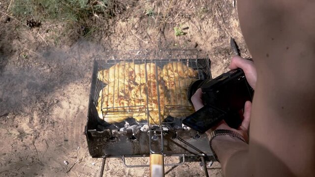 A man films juicy chicken meat grilling over charcoal coals outdoors. Concept of food blogging, culinary content creation, summer barbecue, and outdoor cooking lifestyle.