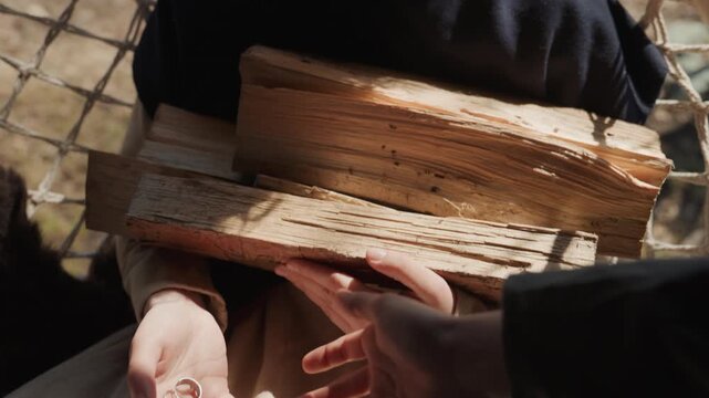 Overhead view shows seated person handling stacked firewood, holding book on lap, scattering seeds from palm, and reaching outward in warm natural light with woven chair texture, calm rustic setting
