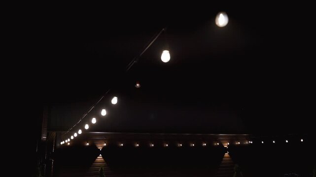 Decorative outdoor string light bulbs hanging on a wire illuminating a dark backyard space and swaying gently in the wind, creating a warm, cozy, and celebratory atmosphere for an evening event