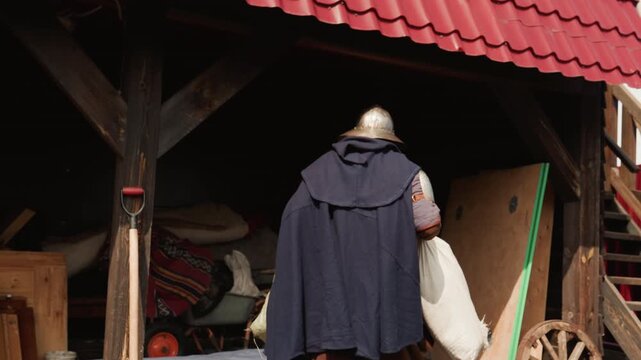 Costumed reenactor with helmet and dark cloak moves through shadowed timber shelter, holds heavy sack at doorway, then continues across rustic village yard beneath soft overcast light