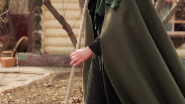 Young woman wearing green wool cloak strides across dry ground while gripping long wooden staff, captured mid-step with one boot lifted and cloak hem swinging to emphasize forward motion and