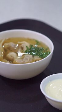 Slow motion camera panning of traditional uzbek chuchvara dumpling soup served with fresh herbs and sour cream, vertical video.