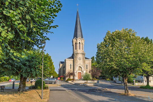 Montrevel-en-Bresse, France: Wide view of the village church with tall steeple framed by lush green trees on a clear sunny day.