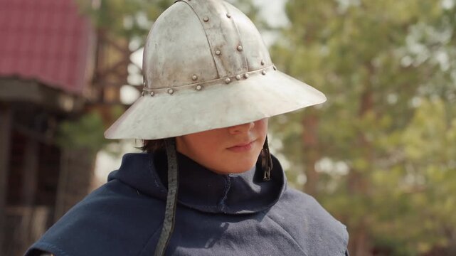 Young medieval reenactor wears weathered kettle helmet, loose chin straps, and padded gambeson while holding calm steady gaze; subtle head turn and slight chin tilt suggest quiet confidence in
