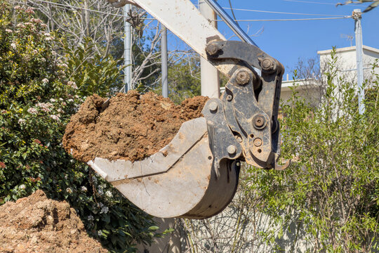 Excavator bucket lifting loose soil during residential garden earthworks under clear blue sky