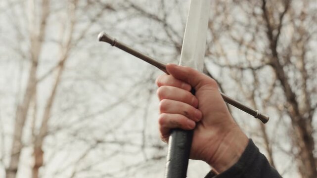 Tight close-up shows hands firmly gripping sword hilt with blade held upright outdoors, tense fingers and wrists against bare trees and cloudy sky, suggesting readiness, inspection, or training