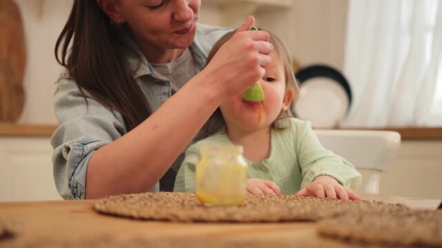 Happy family at home. Mother feeding her baby girl from spoon in kitchen. Little toddler child with messy funny face eats healthy food at home. Young woman mom giving food to kid daughter