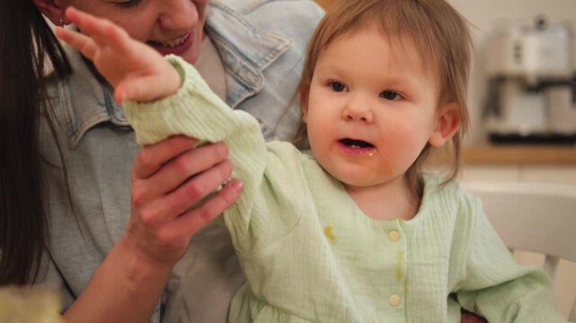 Happy family at home. Mother feeding her baby girl in kitchen. Little toddler child with messy funny face eats healthy food at home. Young woman mom giving food to kid daughter
