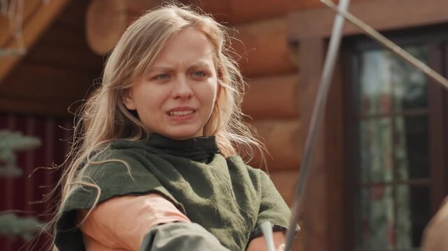 Young woman wearing layered medieval-style clothing holds longbow at chest height beside rustic log cabin, pausing between aim and release with focused expression and slight smile, with archery