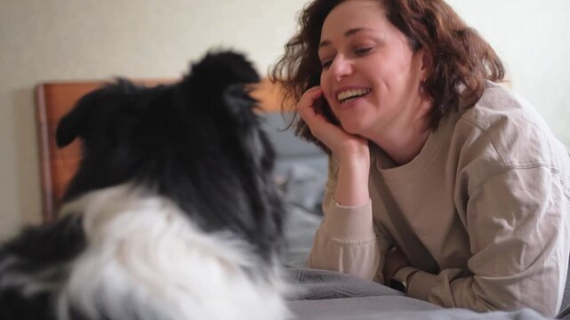 Smiling woman playing with cute puppy dog border collie on bed at home indoor. Owner girl stroking holding dog friend sitting on sofa in living room. Love for pets friendship support team concept