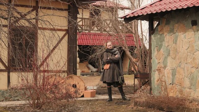 Woman stands paused on cobblestone path in rural courtyard, holding hands with calm contemplative posture; bare shrubs, leafless vines, clay pots, stone walls, timber buildings, and overcast light