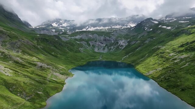 Aerial view of a pristine blue alpine lake nestled in a green mountain valley with snowy peaks under clouds