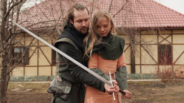Male and female reenactors stand close together in courtyard as male guides female hands along long sword, showing controlled movement and focused expressions during historical training