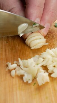 Chef chops fresh garlic cloves on a wooden cutting board with a metal knife. Close-up of a professional preparing aromatic seasoning for a gourmet dish. Vertical video.