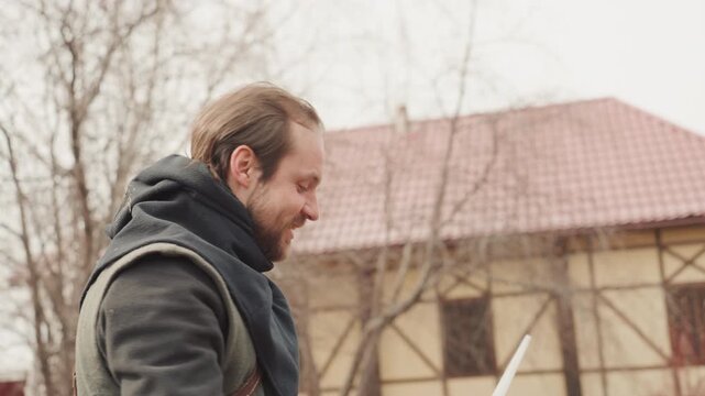 Bearded adult man holds long rod and metal tool outdoors in residential yard, raises hand near eye level, and speaks while inspecting details with focused, practical attention, with man, beard