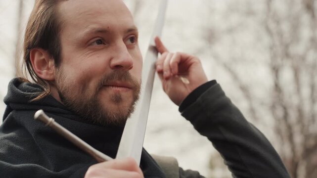 Adult man outdoors holds metal sword across chest near face while inspecting blade and hilt, using careful hand gestures, slight head tilts, and focused gaze to show calm readiness and concentration