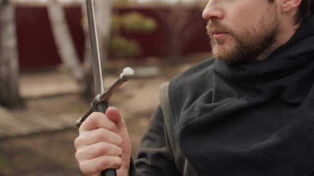 Close-up outdoor scene shows bearded man handling wooden sword with steady grip, checking balance, adjusting hand position, and turning head while studying blade with calm focused expression and