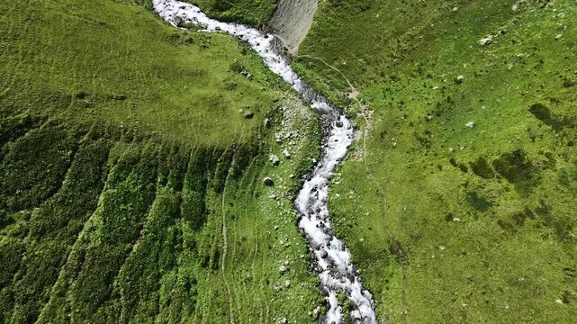 Top-down aerial view of a fast flowing mountain river cutting through green alpine meadows